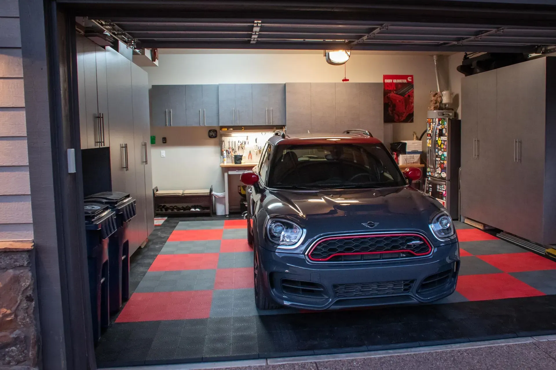 A dark gray Mini Cooper parked in a garage with a red and gray checkered floor, organized cabinets, and storage units.