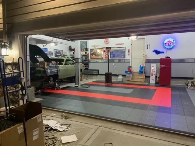 A garage with a car on a lift, a tiled gray and red floor, a red refrigerator, and a neon sign on the back wall.