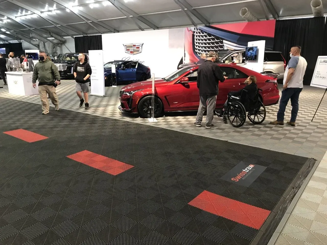 People walk past a red sedan displayed at a Cadillac event in an indoor exhibit hall with patterned black floor mats.