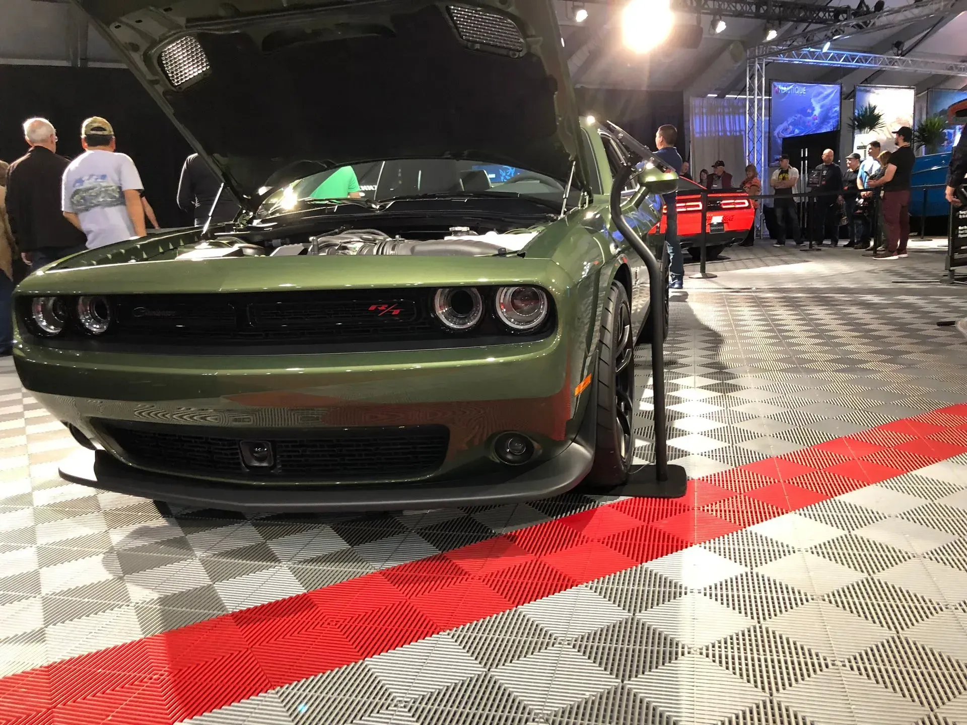 A green Dodge Challenger with its hood open, displayed on a checkered floor at an indoor car show with people nearby.