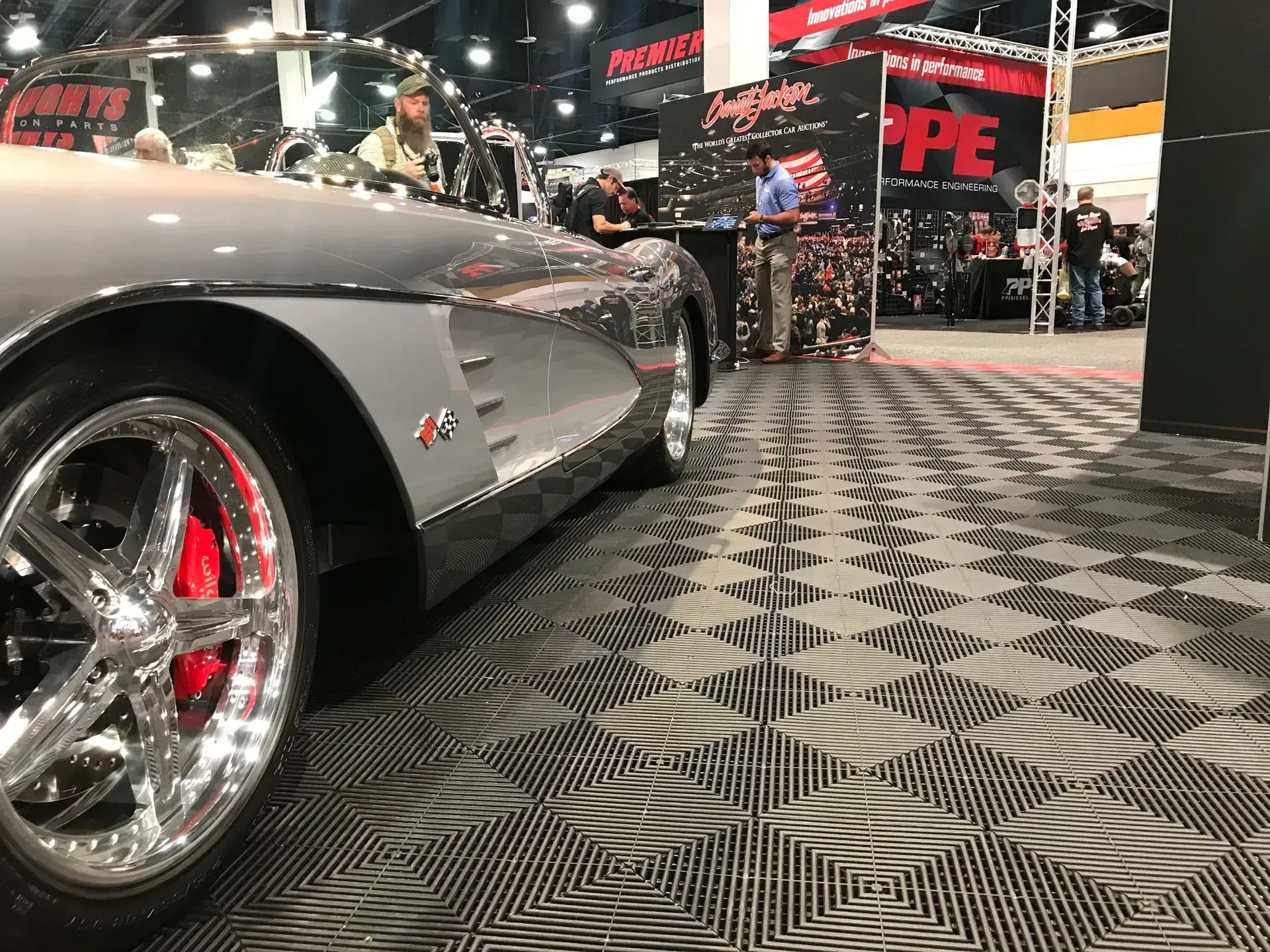 A grey classic convertible Corvette on display at an indoor trade show with black and grey tiled flooring.