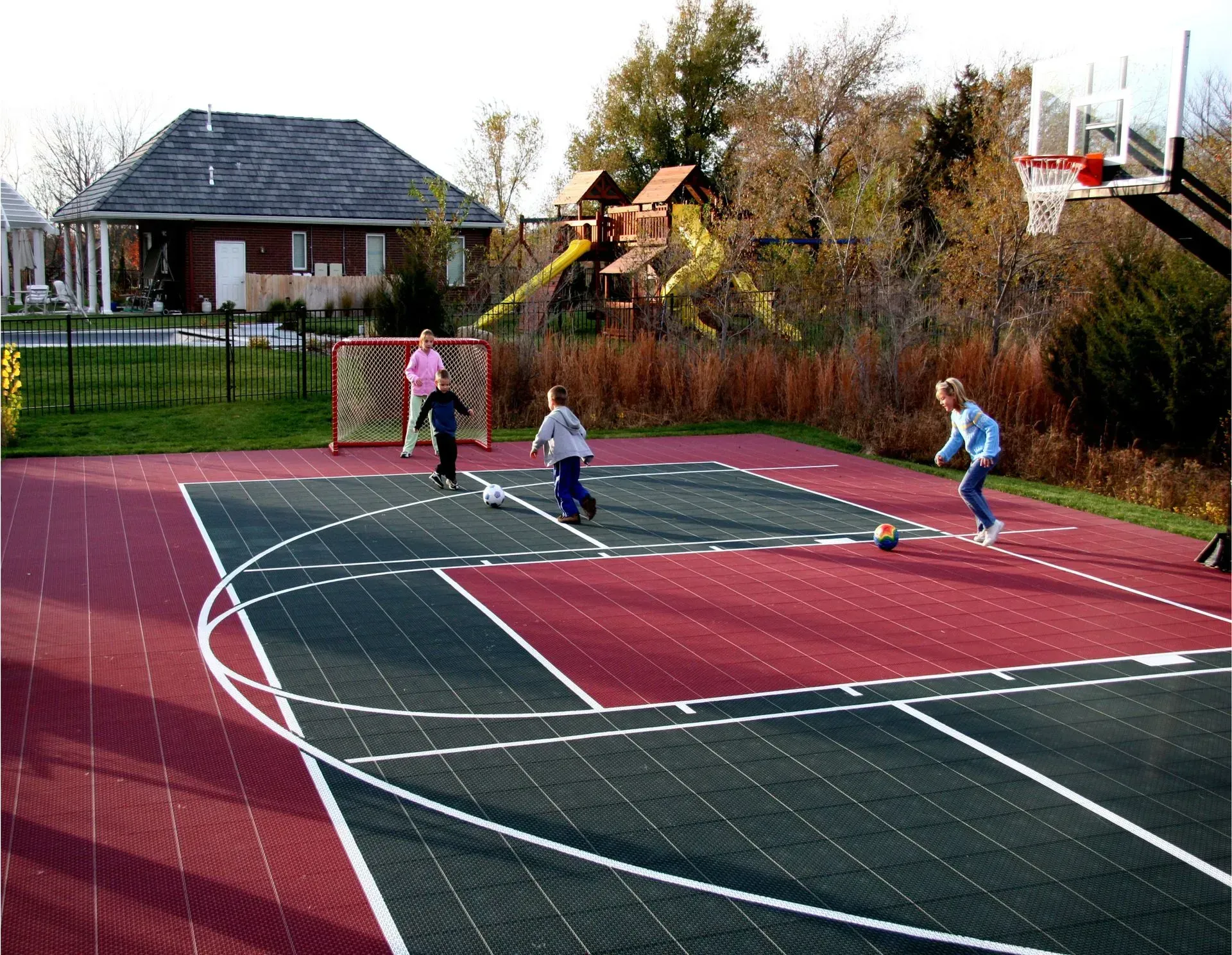Four children play with a soccer ball on a red and green outdoor sports court near a house and a playset.