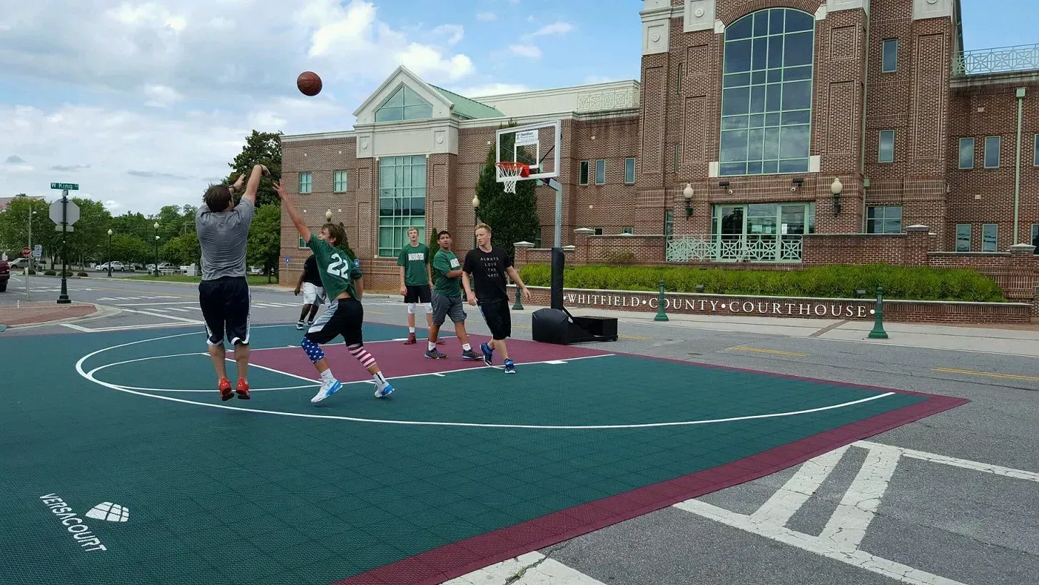 People play basketball on a blue and purple outdoor court in front of a brick building on a sunny day.