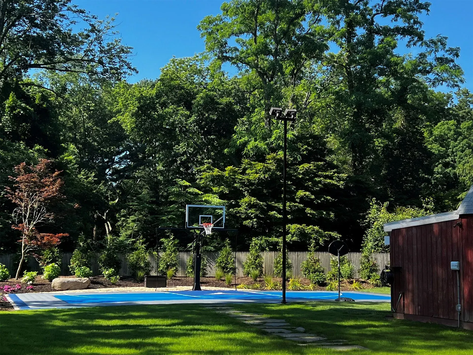 A blue outdoor basketball court set in a grassy backyard surrounded by lush green trees and a red wooden structure.