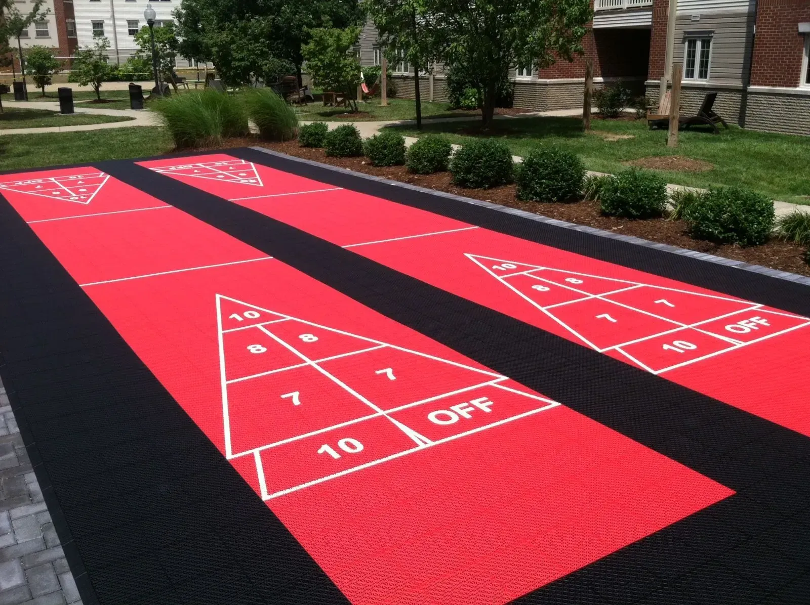 Two red outdoor shuffleboard courts with black borders and white markings, set on a lawn near a brick building.