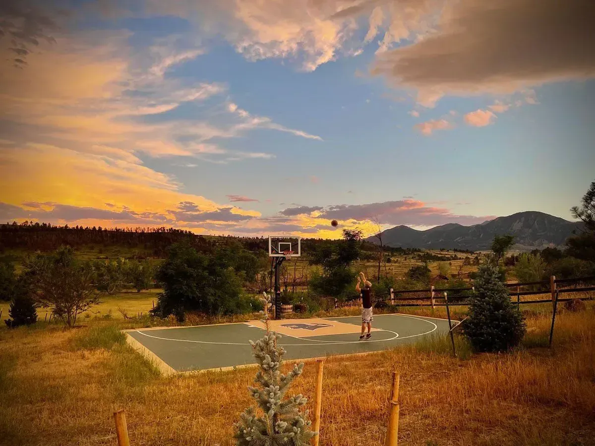 Two people playing on an outdoor basketball court at sunset, set against a backdrop of mountains and a colorful sky.