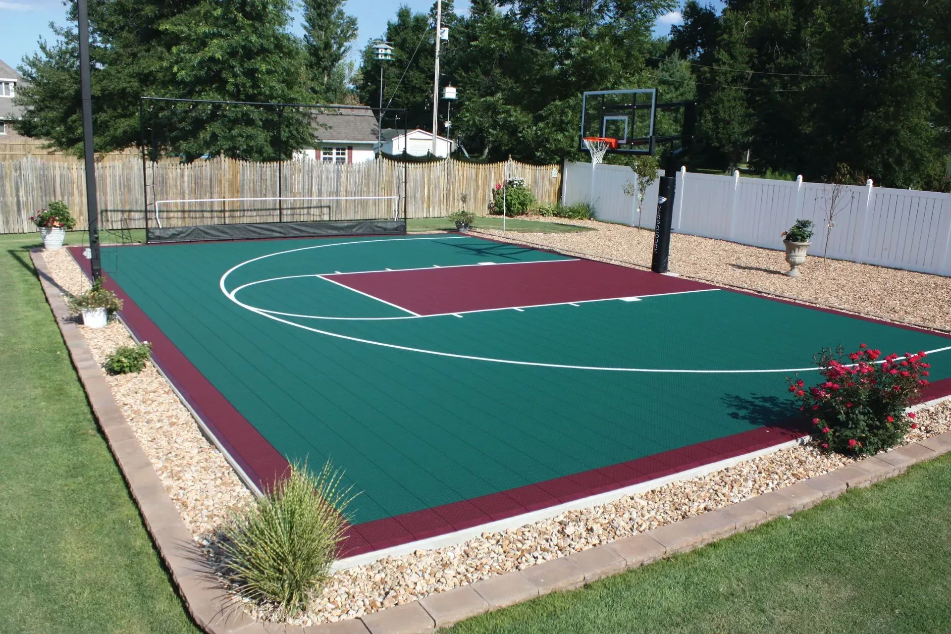 A backyard basketball court with a green surface, maroon key, and white hoop, surrounded by gravel and a white fence.