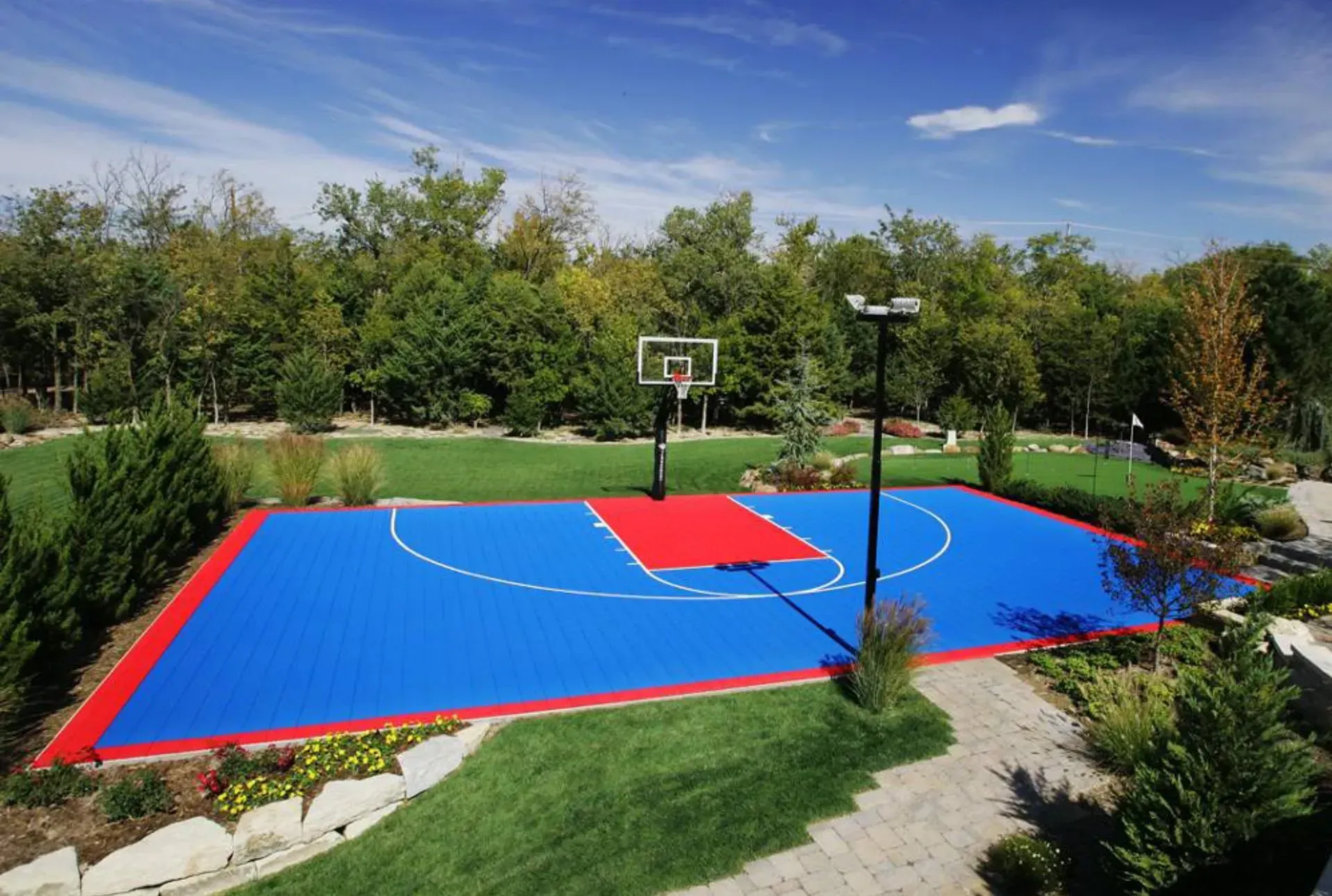 An outdoor basketball court with a blue surface, a red key, white court lines, and a hoop set against a backdrop of trees.