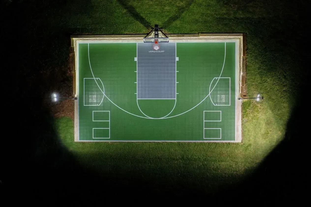 An aerial view of a brightly lit, green basketball court surrounded by dark grass at night.