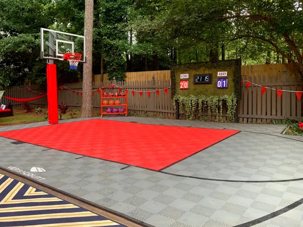 An outdoor backyard basketball court with a red center, gray perimeter, a basketball hoop, and a digital scoreboard.