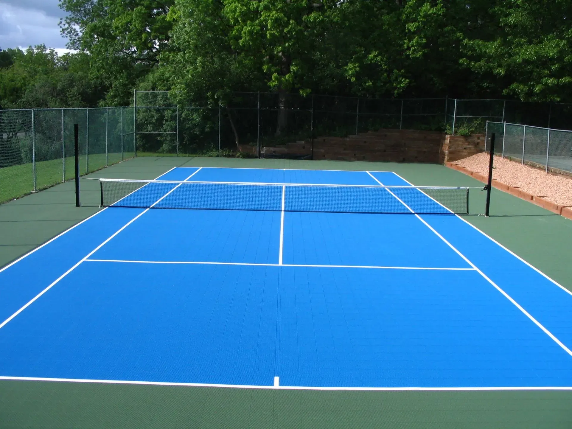 A bright blue tennis court with a center net, surrounded by a fence and trees on a sunny day.