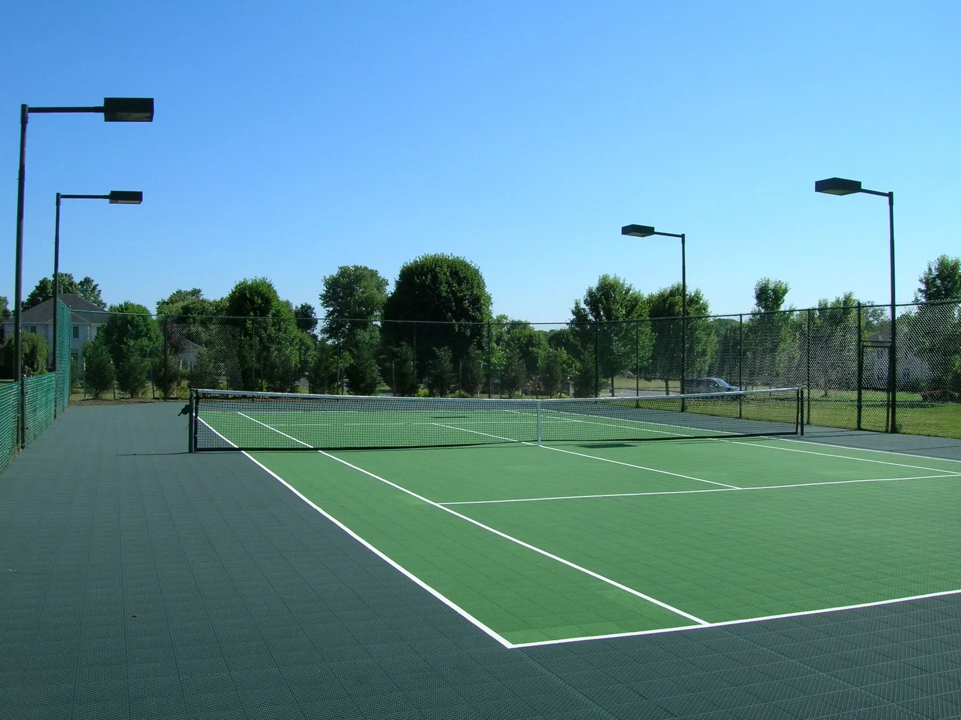 An outdoor tennis court with a green surface, white boundary lines, a central net, and tall light poles against a blue sky.