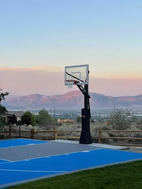 A basketball hoop stands on a blue and gray court outdoors, set against a background of mountains at sunset.