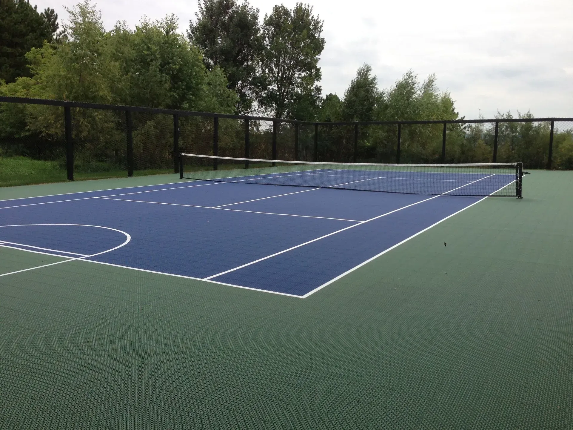 A dual-colored blue and green tennis court with white boundary lines, surrounded by a black fence and trees.