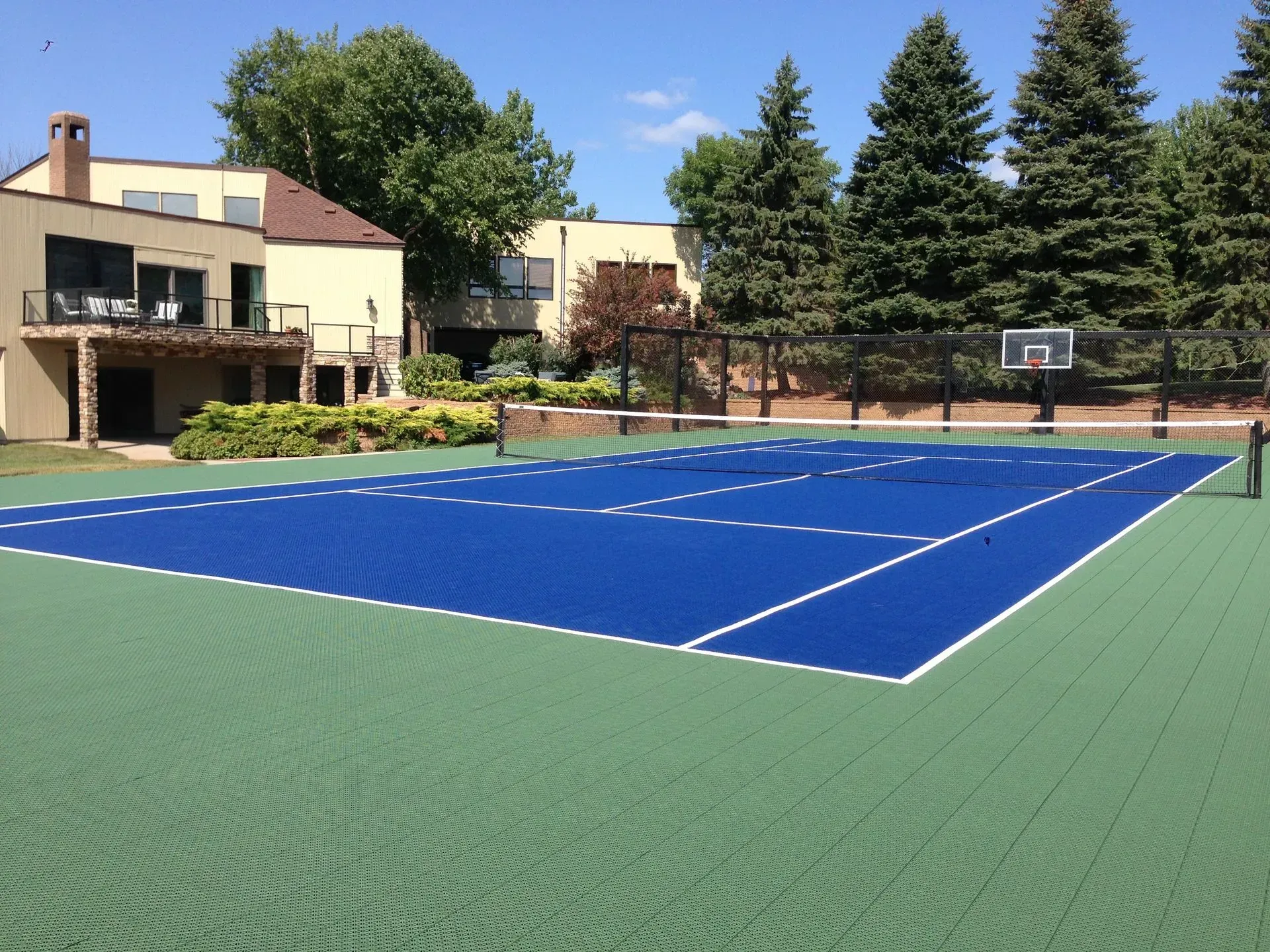 A multi-use outdoor sports court with a blue center tennis area and green perimeter, next to a house and evergreen trees.