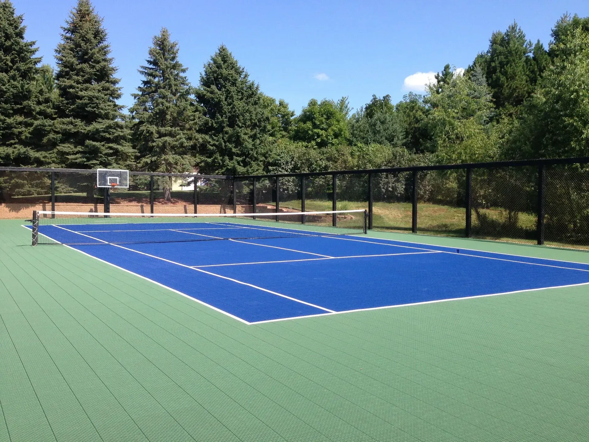 A blue sport court with white lines and a tennis net, surrounded by a black fence and trees.