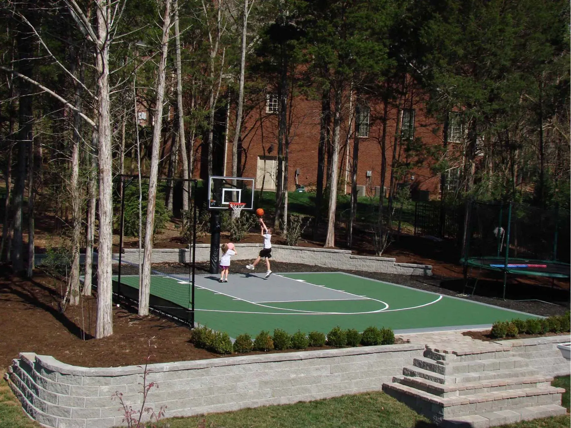 Two people play basketball on a backyard court with green and gray surfacing, surrounded by a stone wall and trees.