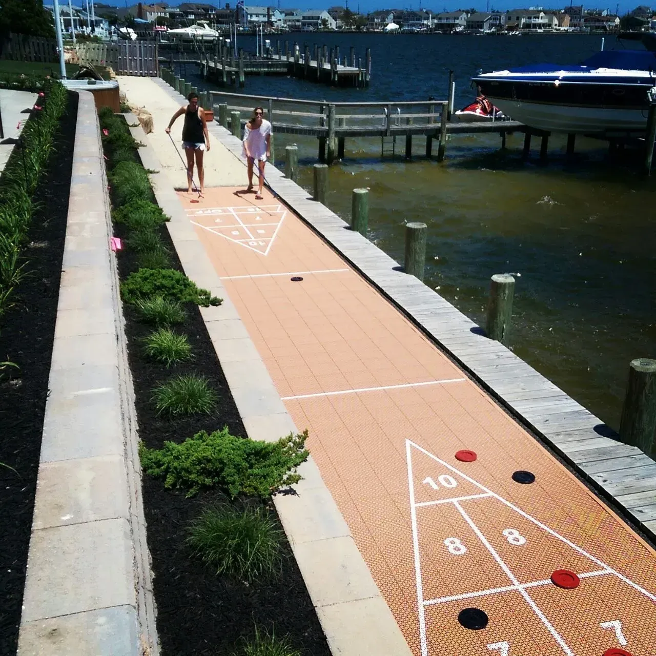 Two people play shuffleboard on an outdoor court overlooking a waterfront with boats and docks.