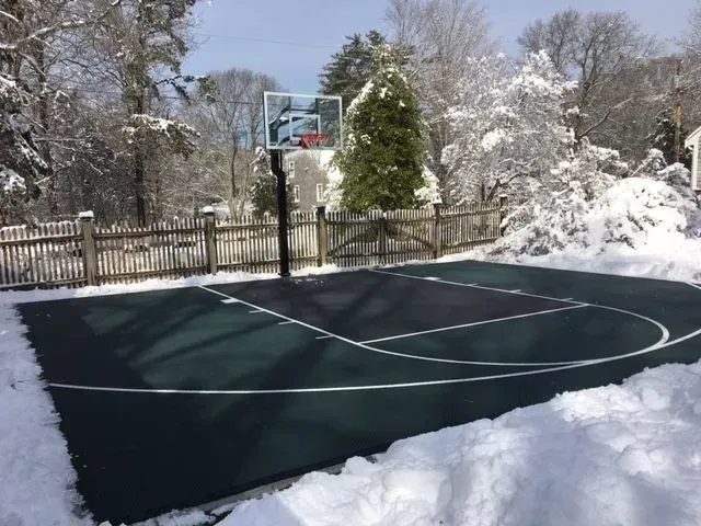 A green and black basketball court surrounded by snow-covered trees and a wooden fence on a sunny winter day.