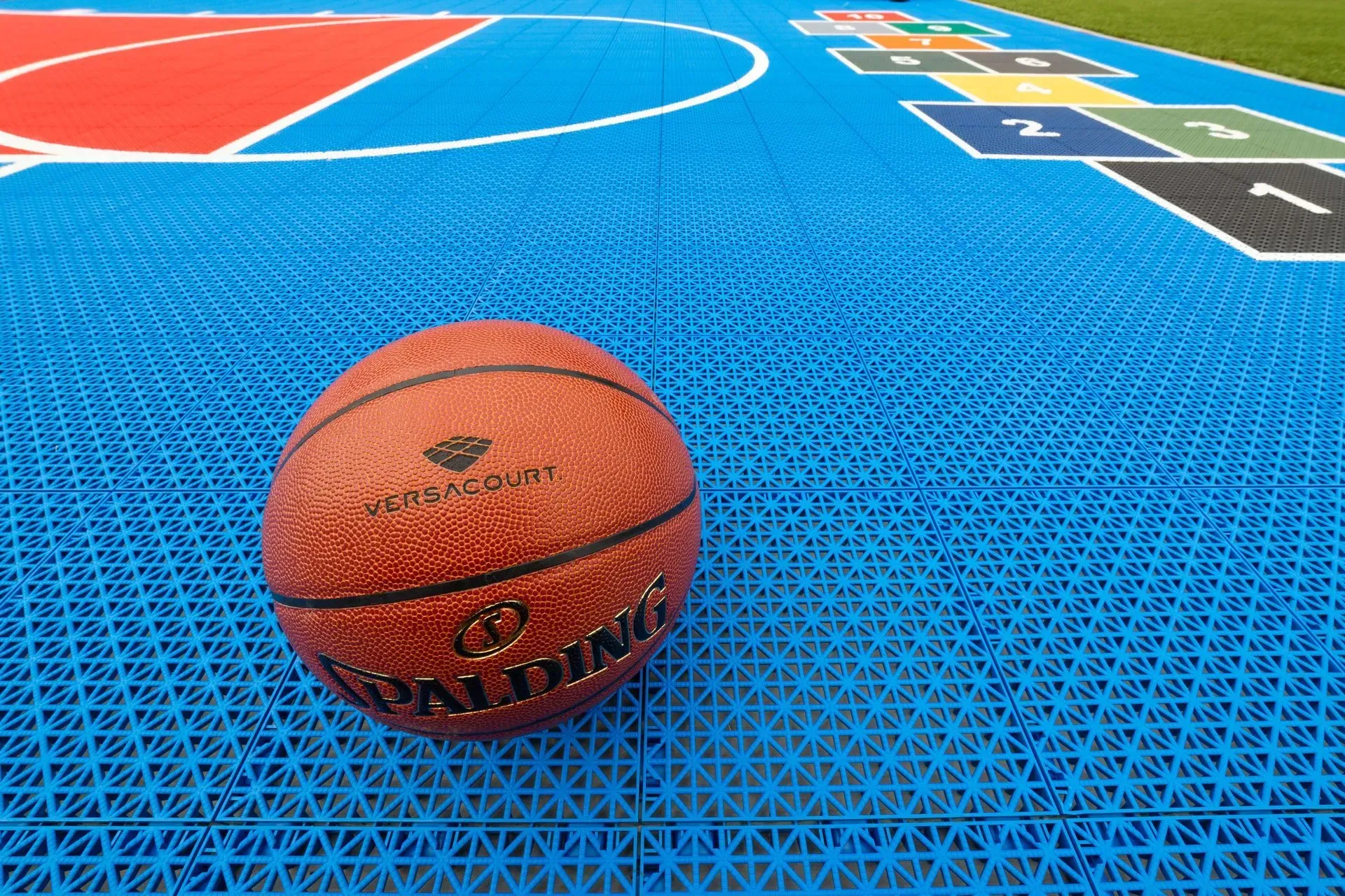 A Spalding basketball rests on a blue outdoor court featuring a red-painted area and a hopscotch grid.