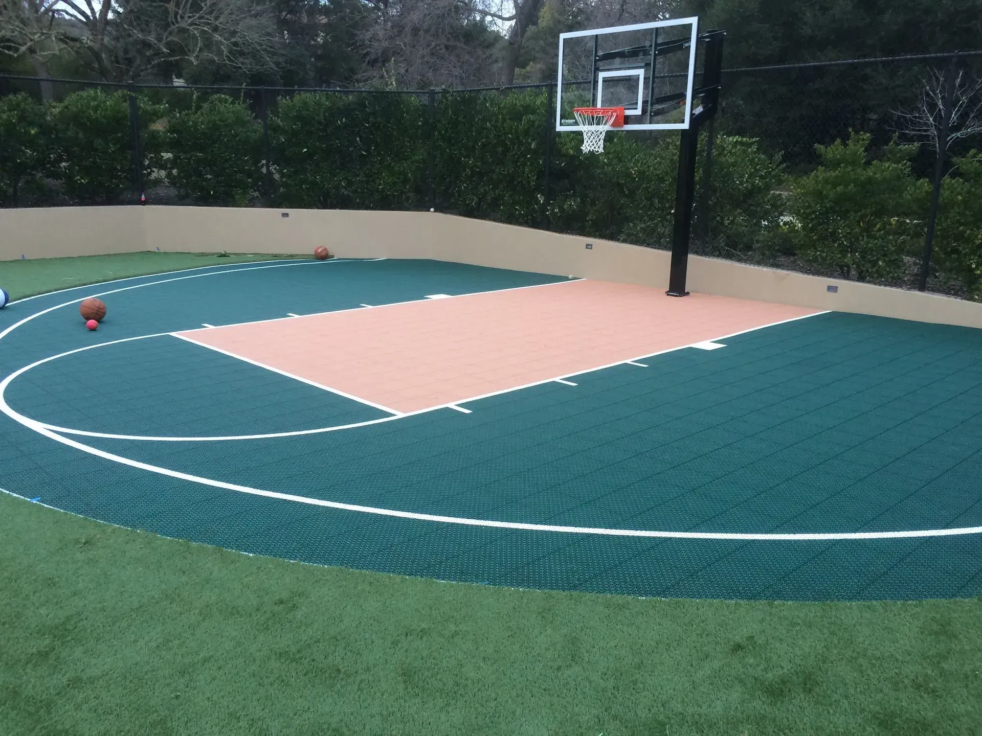 A basketball court with a green surface, a tan key, and a hoop, surrounded by a low wall and lawn in a yard.
