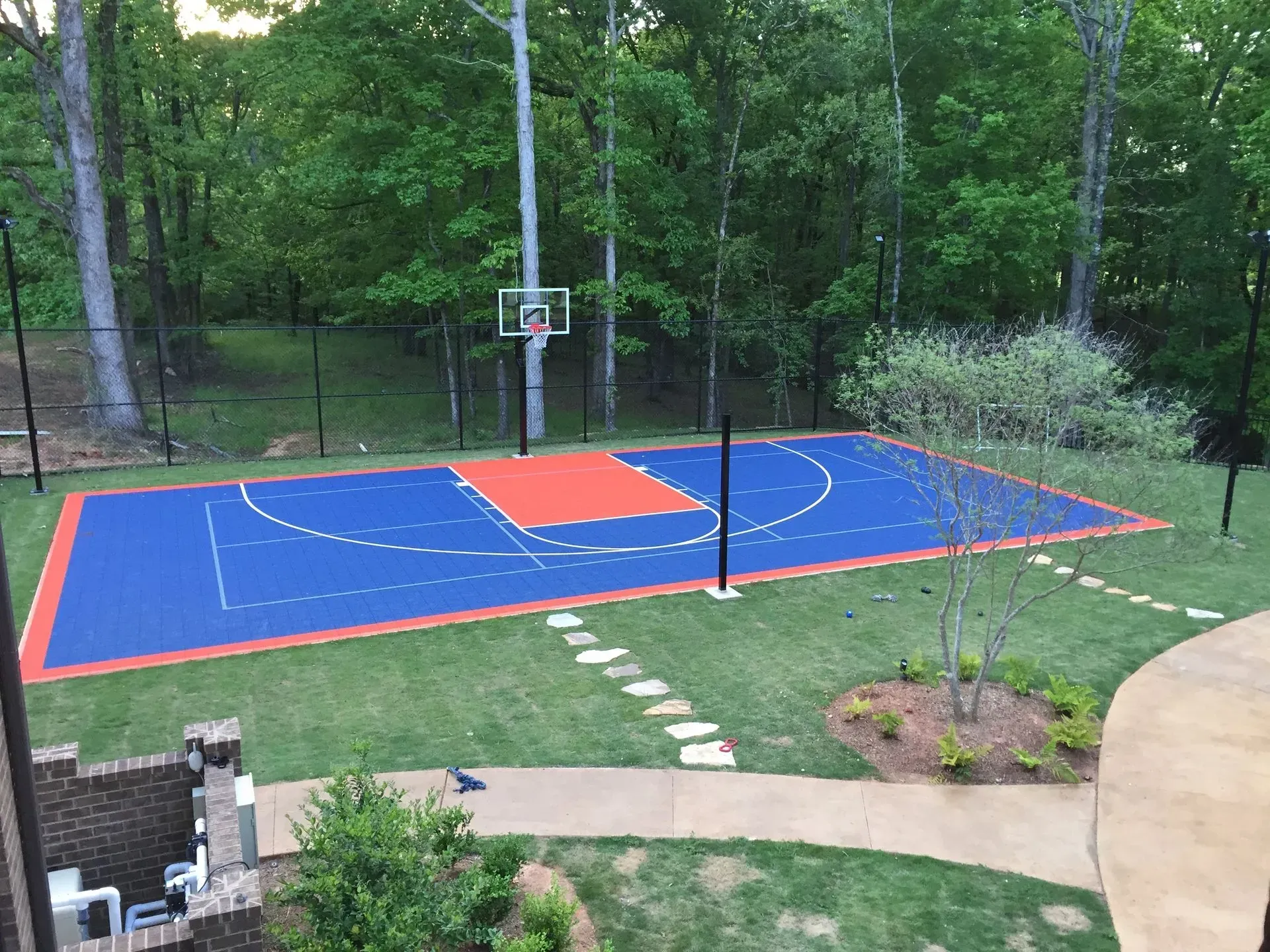 A backyard basketball court with a blue surface, orange key, and white court lines, surrounded by a black fence and trees.