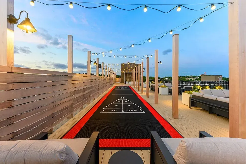 A rooftop deck at dusk featuring a shuffleboard court, string lights, and patio seating.