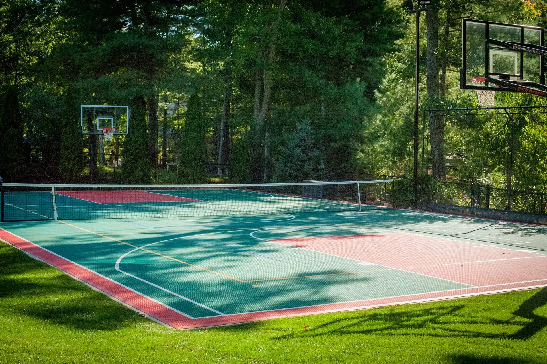 A multi-use outdoor sports court with basketball hoops and a tennis net, featuring teal and red tiles in a grassy area.