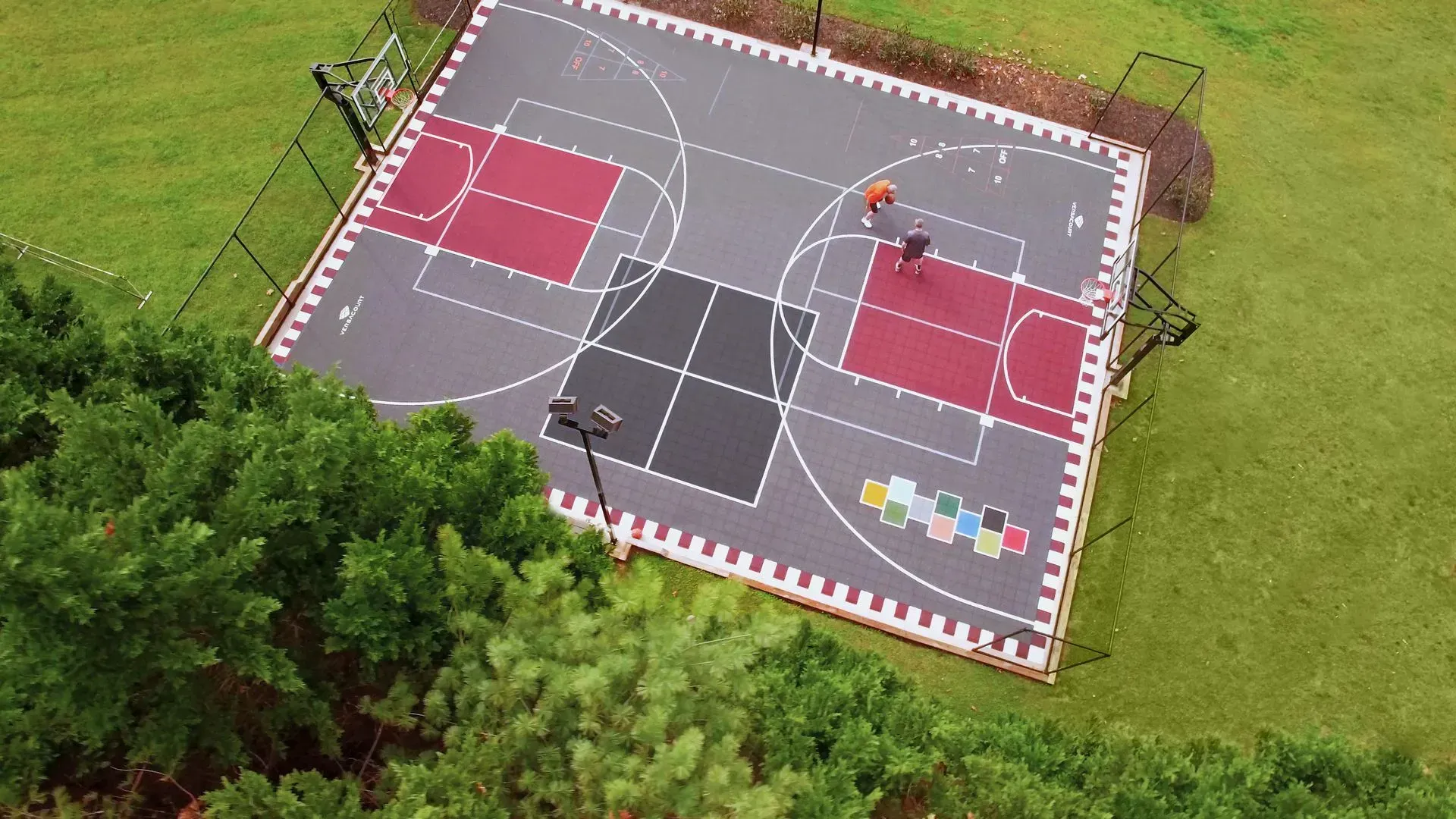 An aerial view of a multi-purpose sports court with two red key areas and a central black square set in a grassy yard.