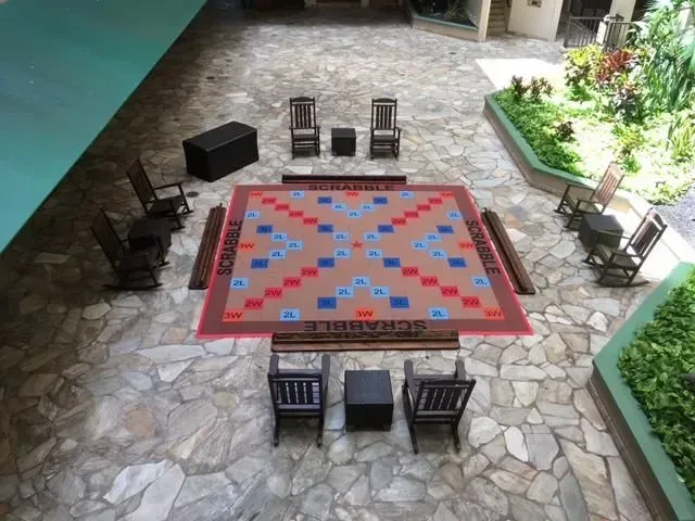 A large, outdoor Scrabble board rug laid out on a stone patio surrounded by wooden rocking chairs and small tables.