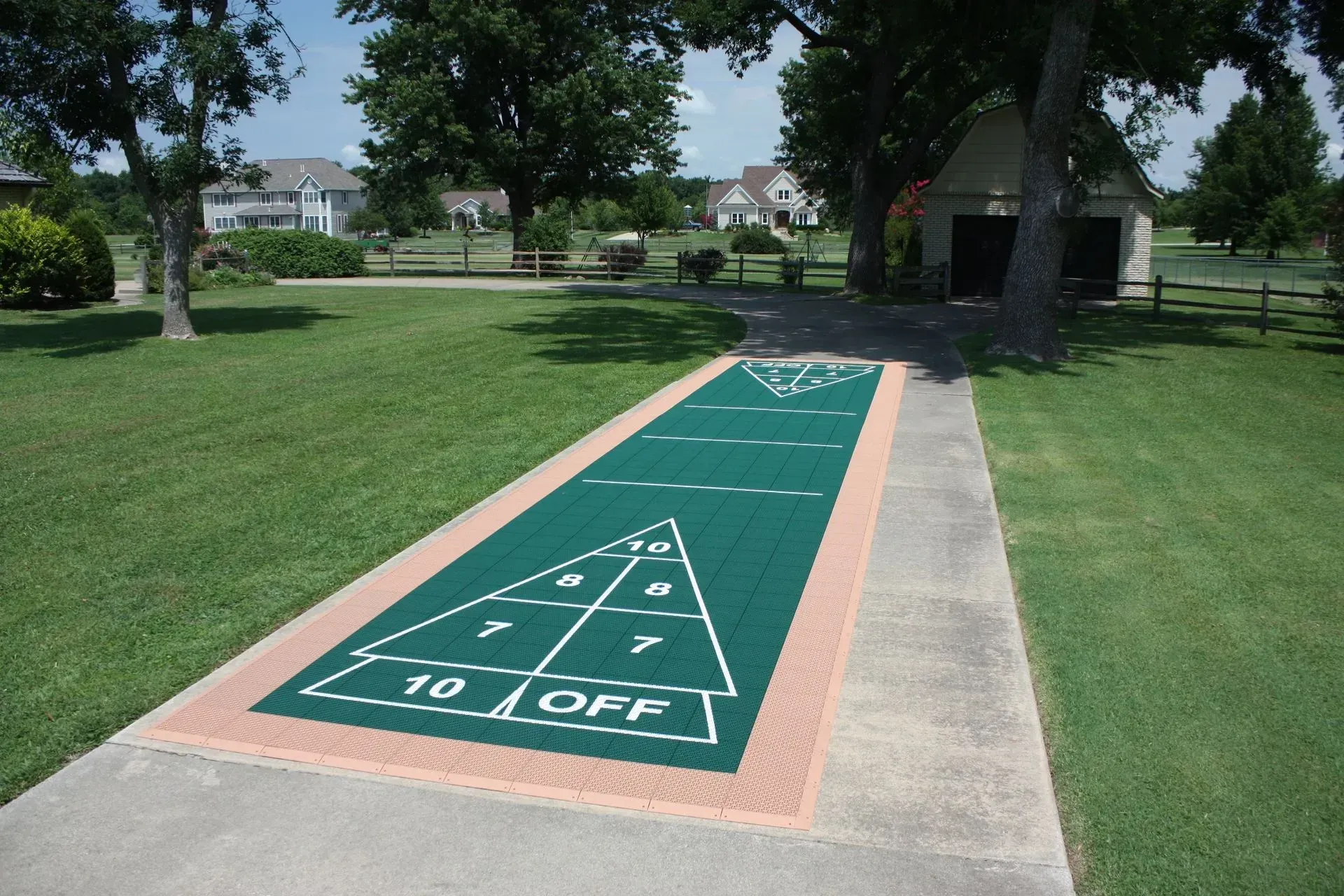 An outdoor shuffleboard court with a green surface and white markings, set on a concrete path in a grassy yard.