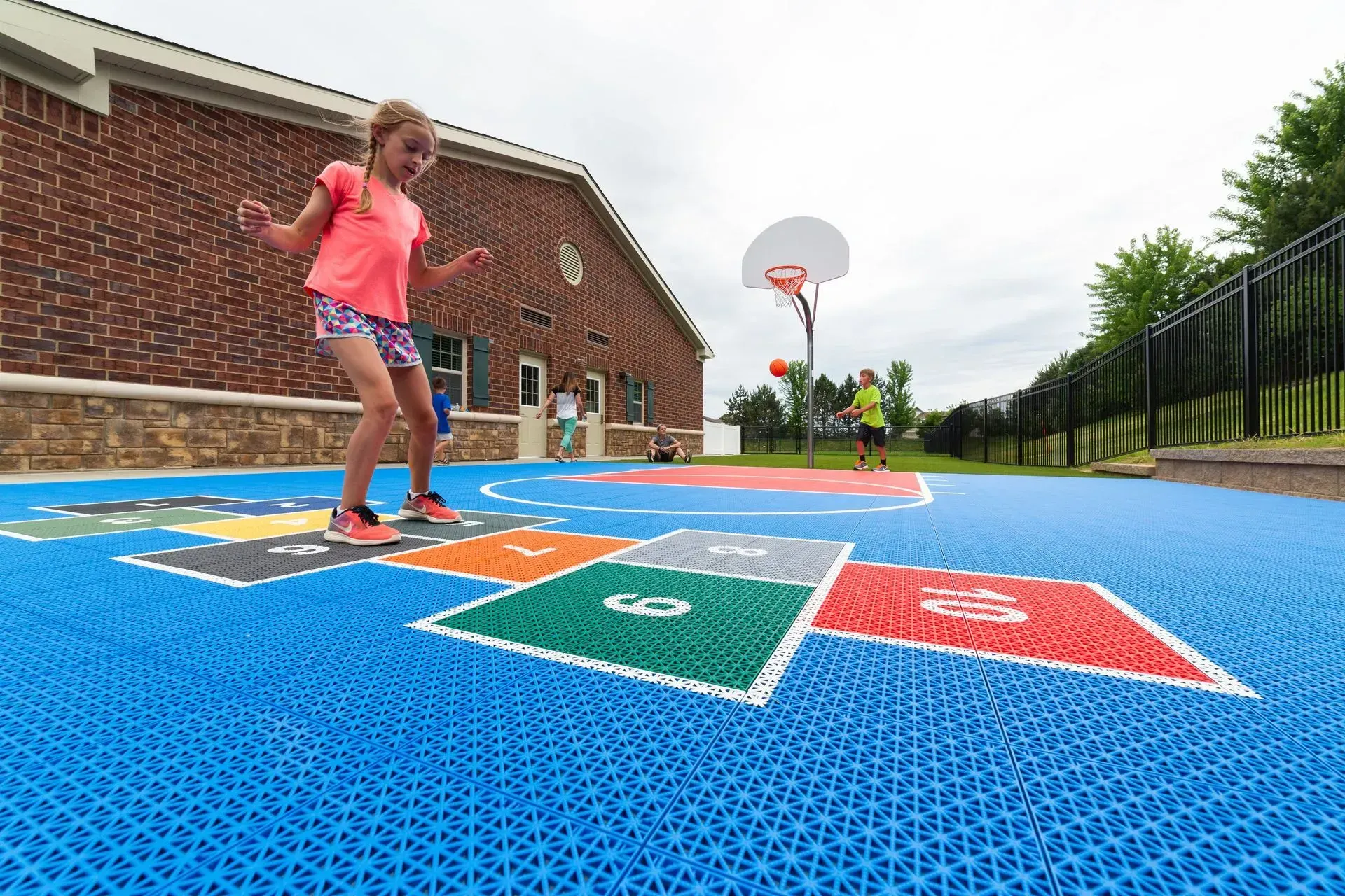A child plays hopscotch on a bright blue outdoor court near a brick building with a basketball hoop in the background.
