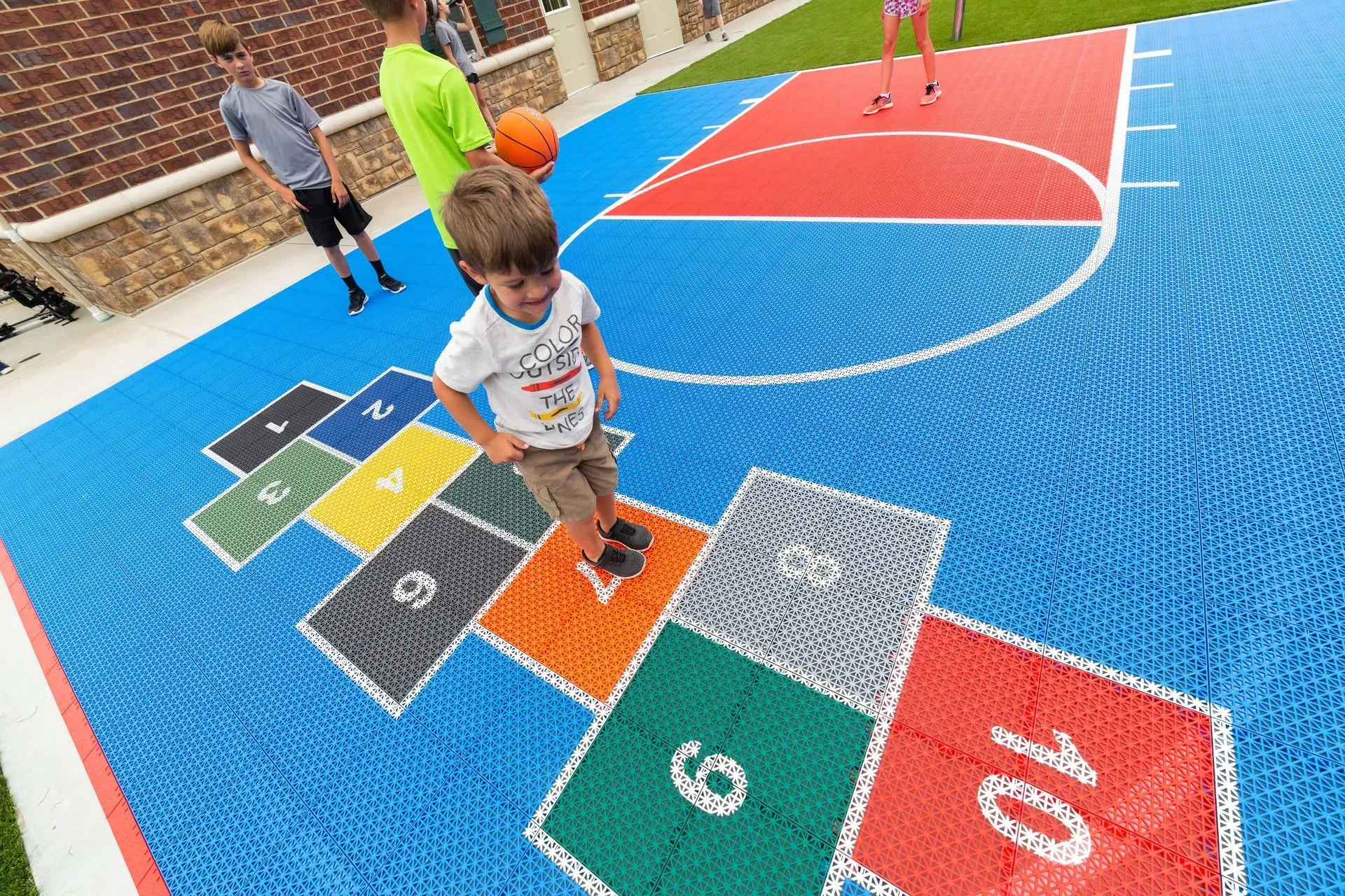 A child playing hopscotch on a colorful, outdoor multi-sport court with others standing in the background.