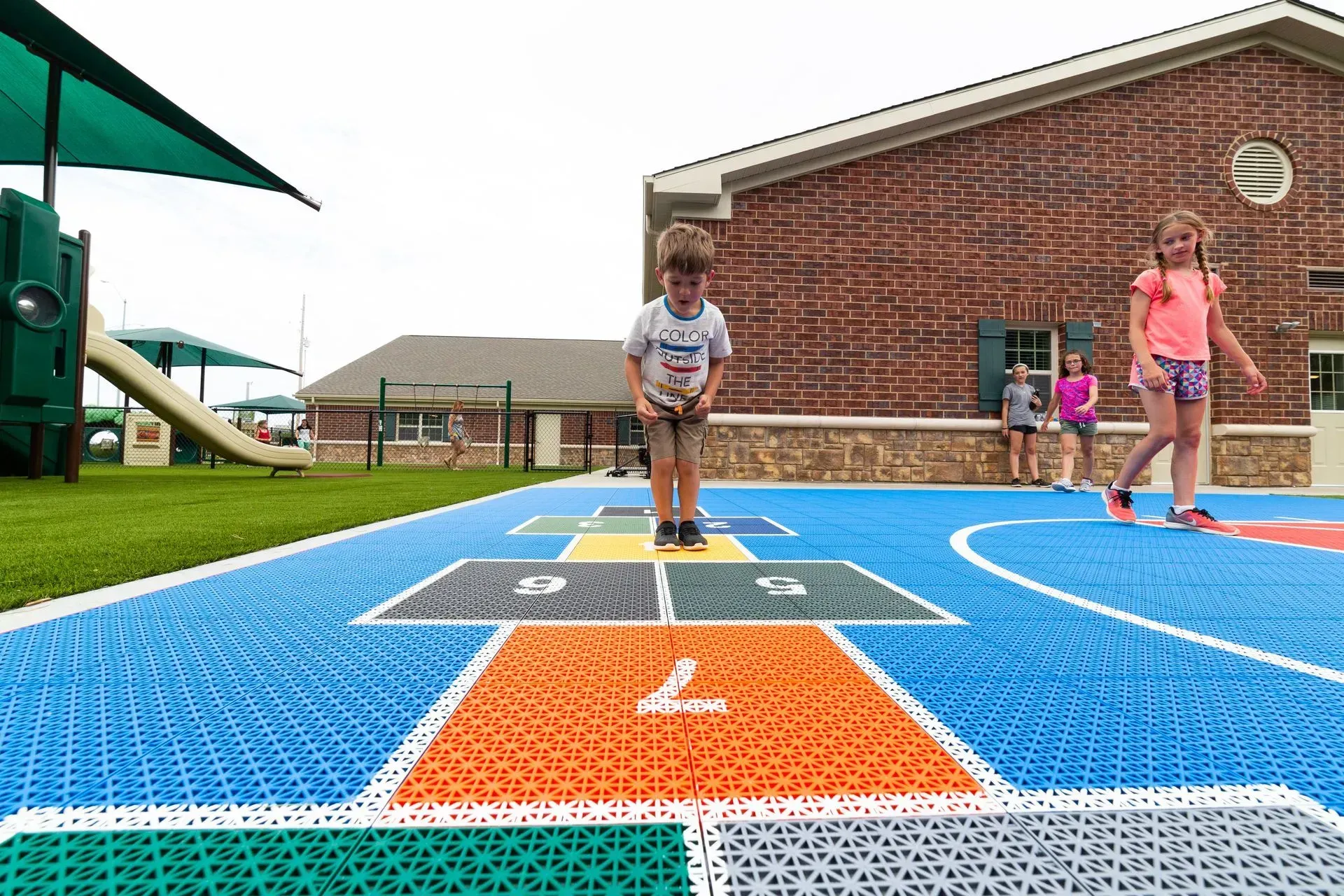 Children play on a colorful, outdoor hopscotch court with a brick building in the background.