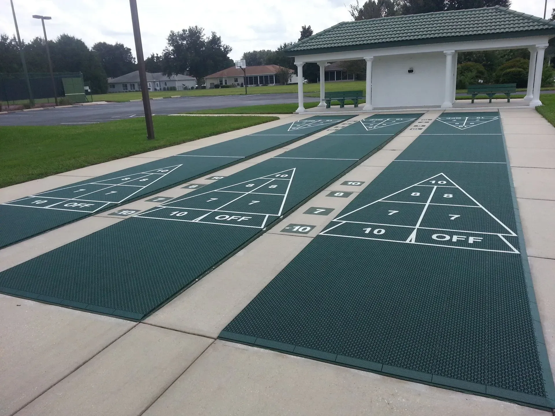 Three green shuffleboard courts with white markings outdoors, adjacent to a small covered seating pavilion.