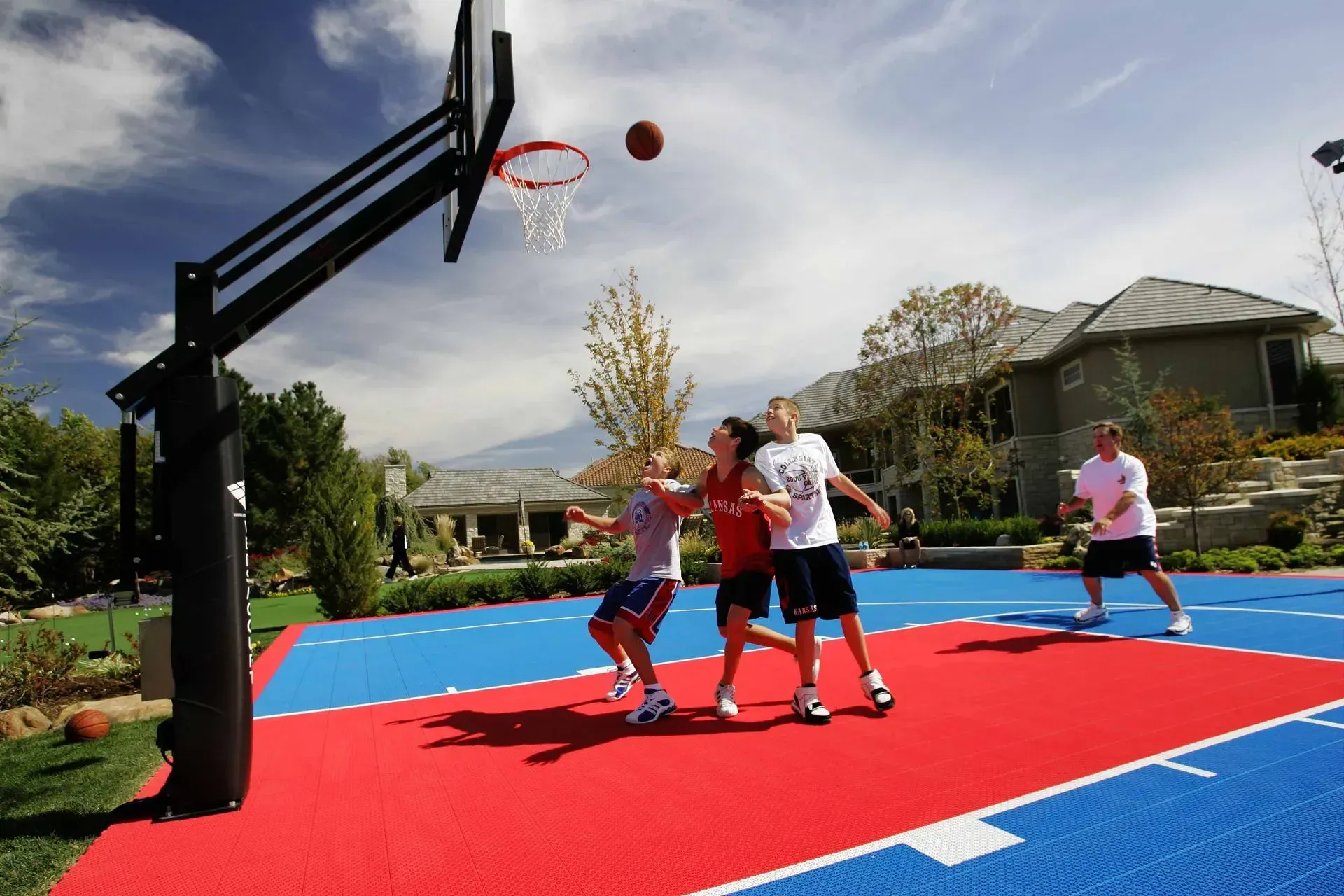 Four people play basketball on a brightly colored red and blue outdoor court near a house under a clear, sunny sky.