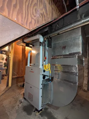 Gas furnace beside ductwork in a basement or utility room, with exposed pipes and insulation.