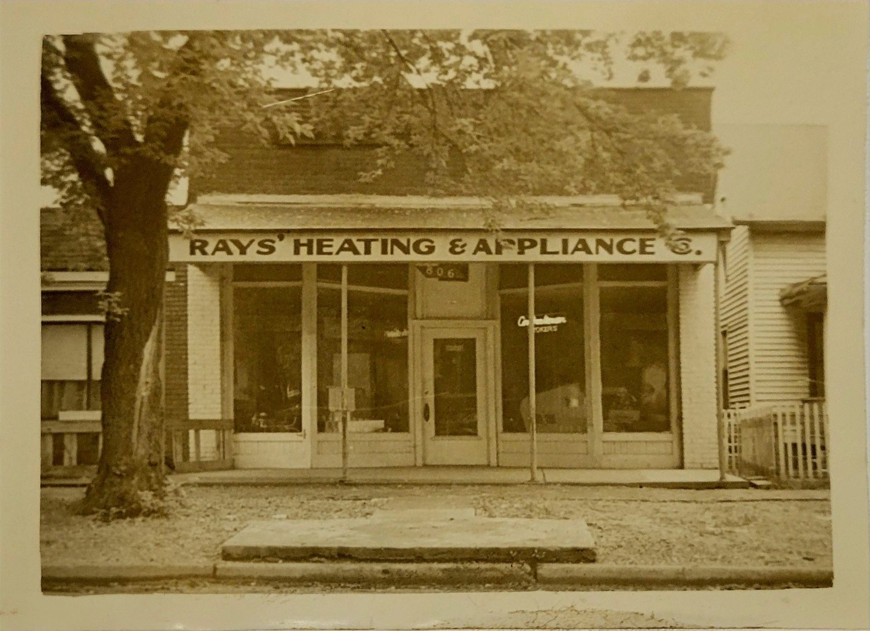 Ray's Heating & Appliance storefront. Window display, signage, tree, and bench in front.