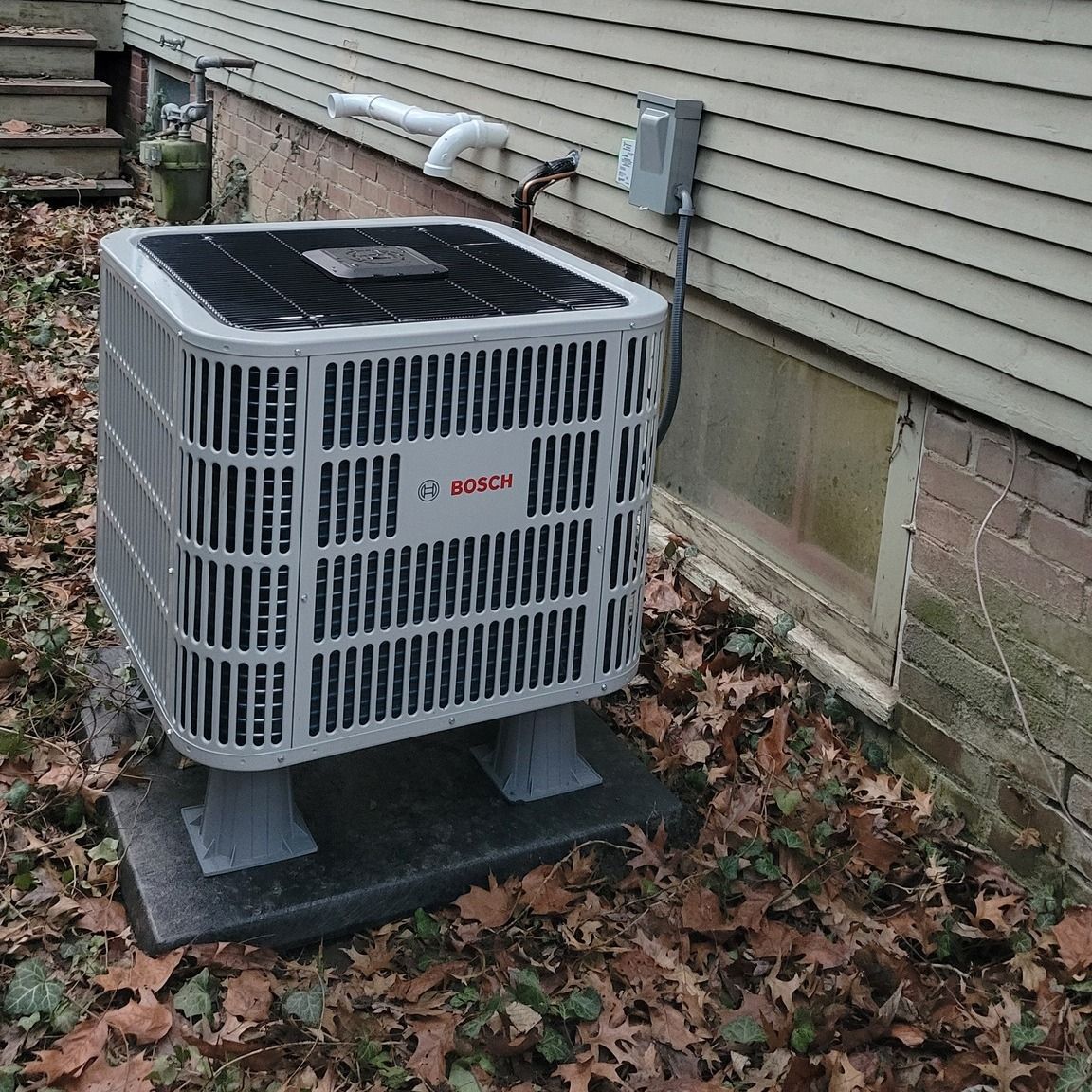 Bosch air conditioning unit outside a building on a concrete base, next to a window.