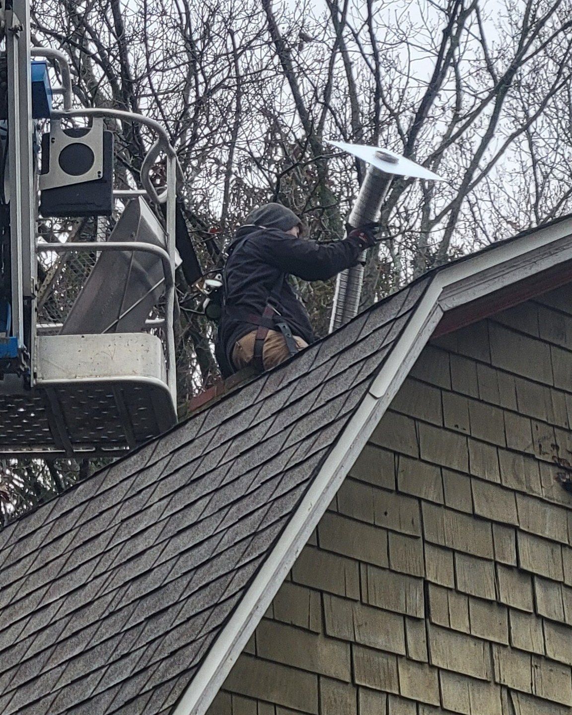Person on roof, installing metal chimney cap, aided by lift; overcast day.