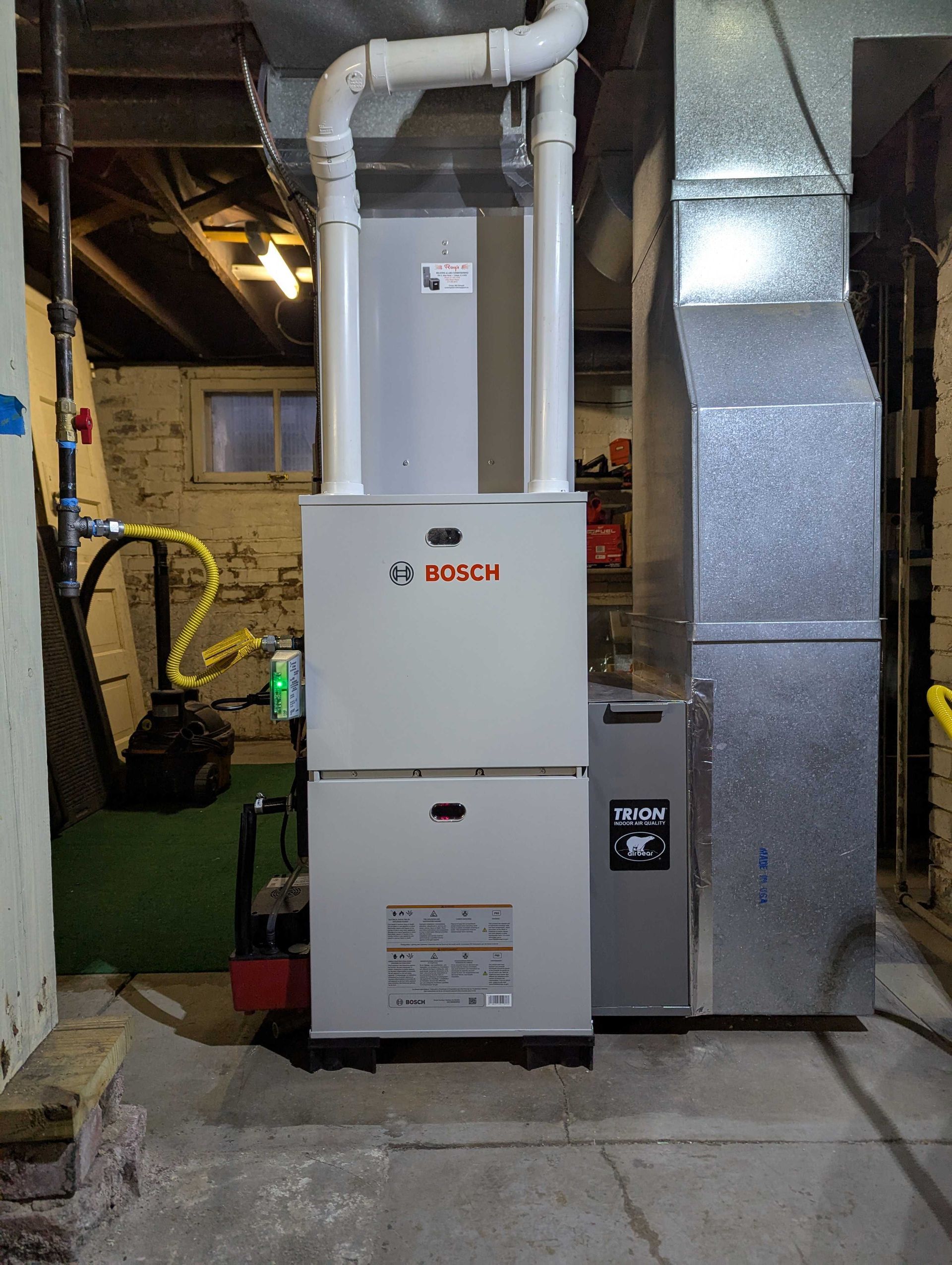 Bosch furnace in a basement. Gray furnace with white vents and ductwork. Concrete floor.