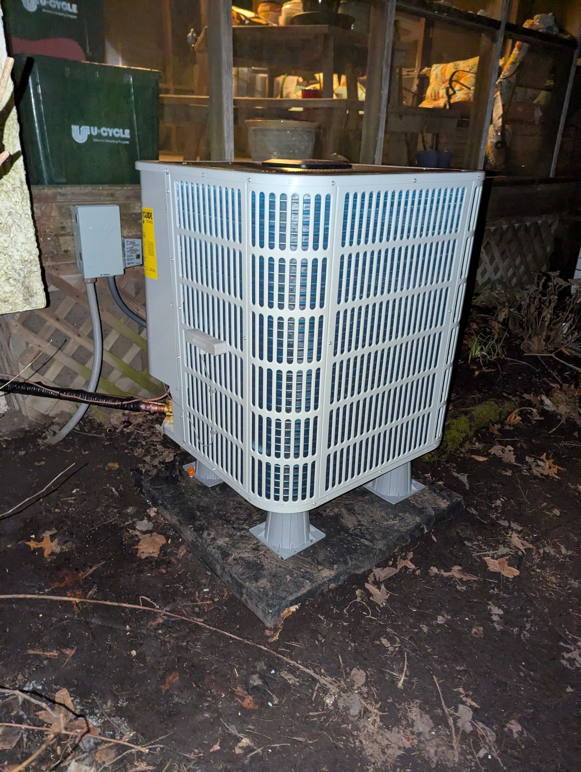 An outdoor air conditioning unit is against a glass structure, on a muddy surface.