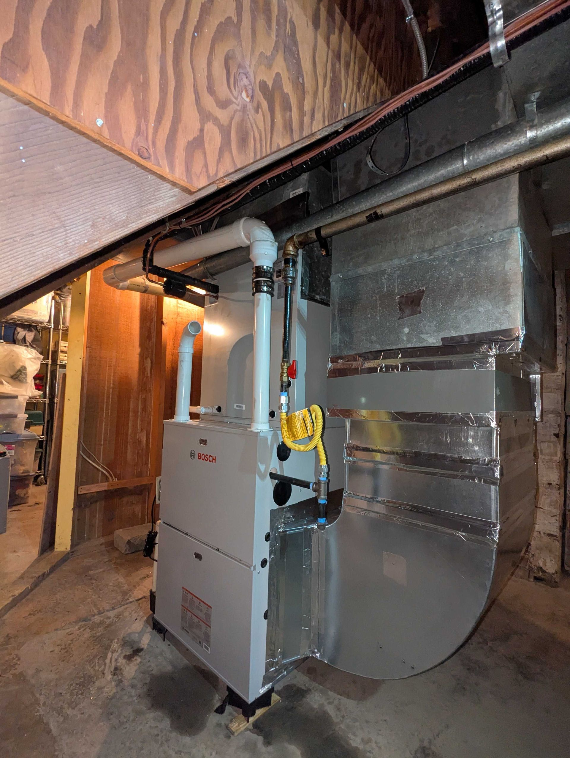 Furnace in a basement. Gray metal unit with white pipes and ductwork. Wooden beams above.