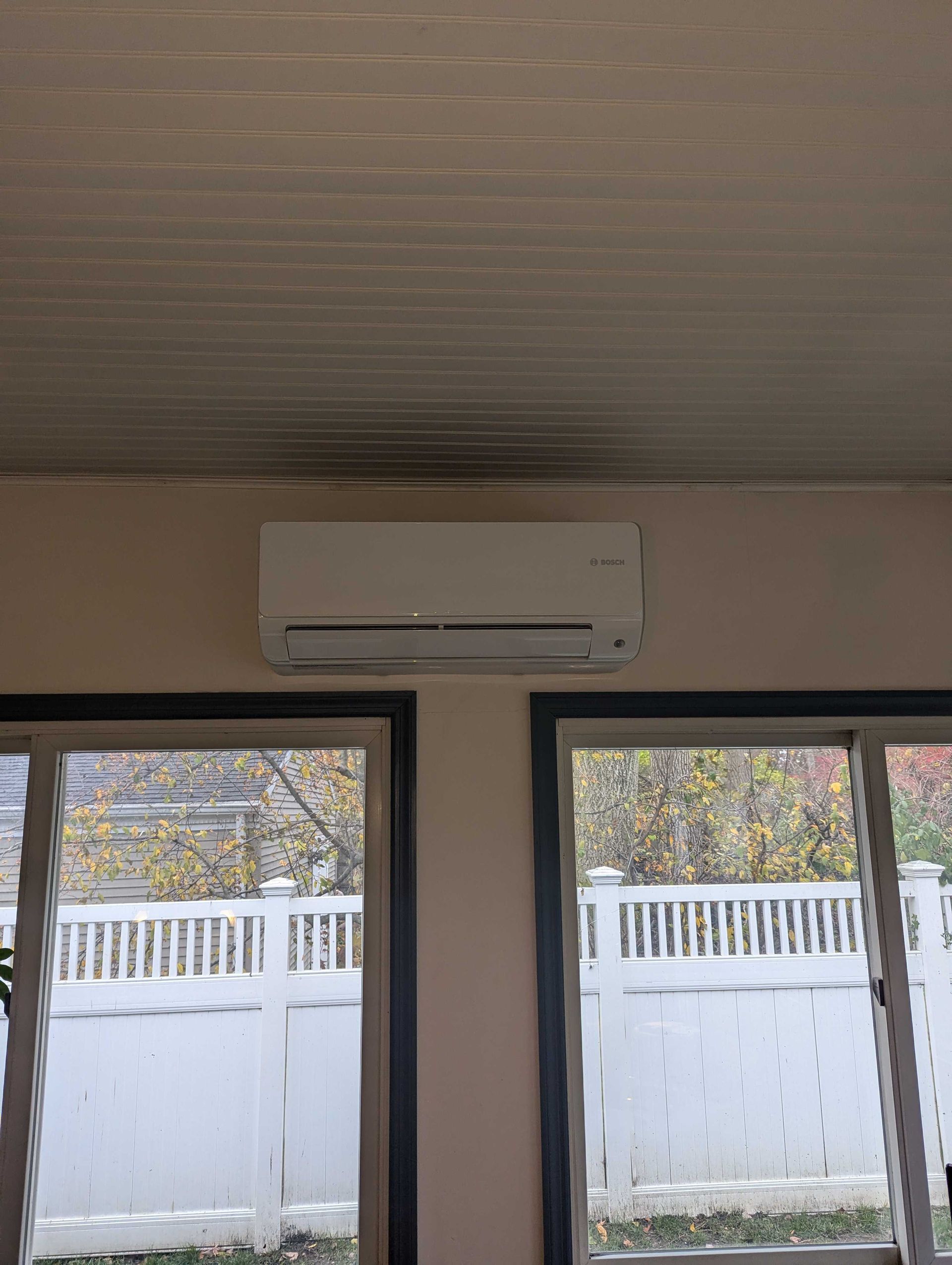 White ductless mini-split air conditioner above two windows with a white fence in the background.