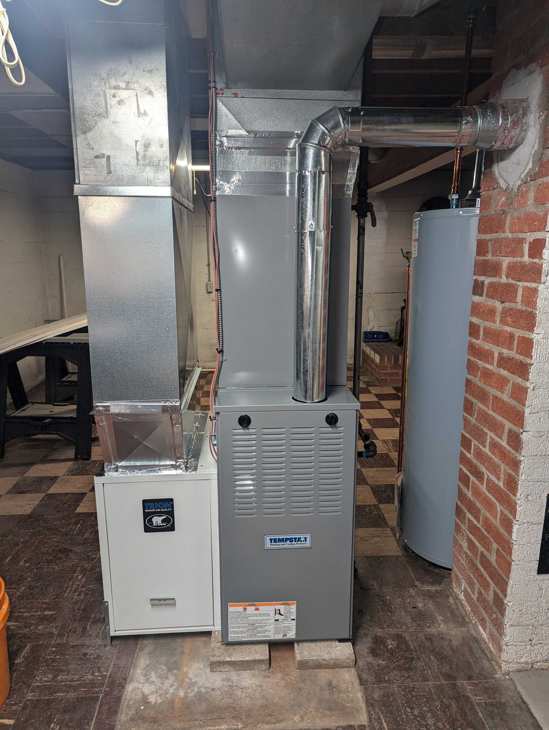 Furnace and air filter system in a basement setting; silver metal and gray units, wooden beams above.