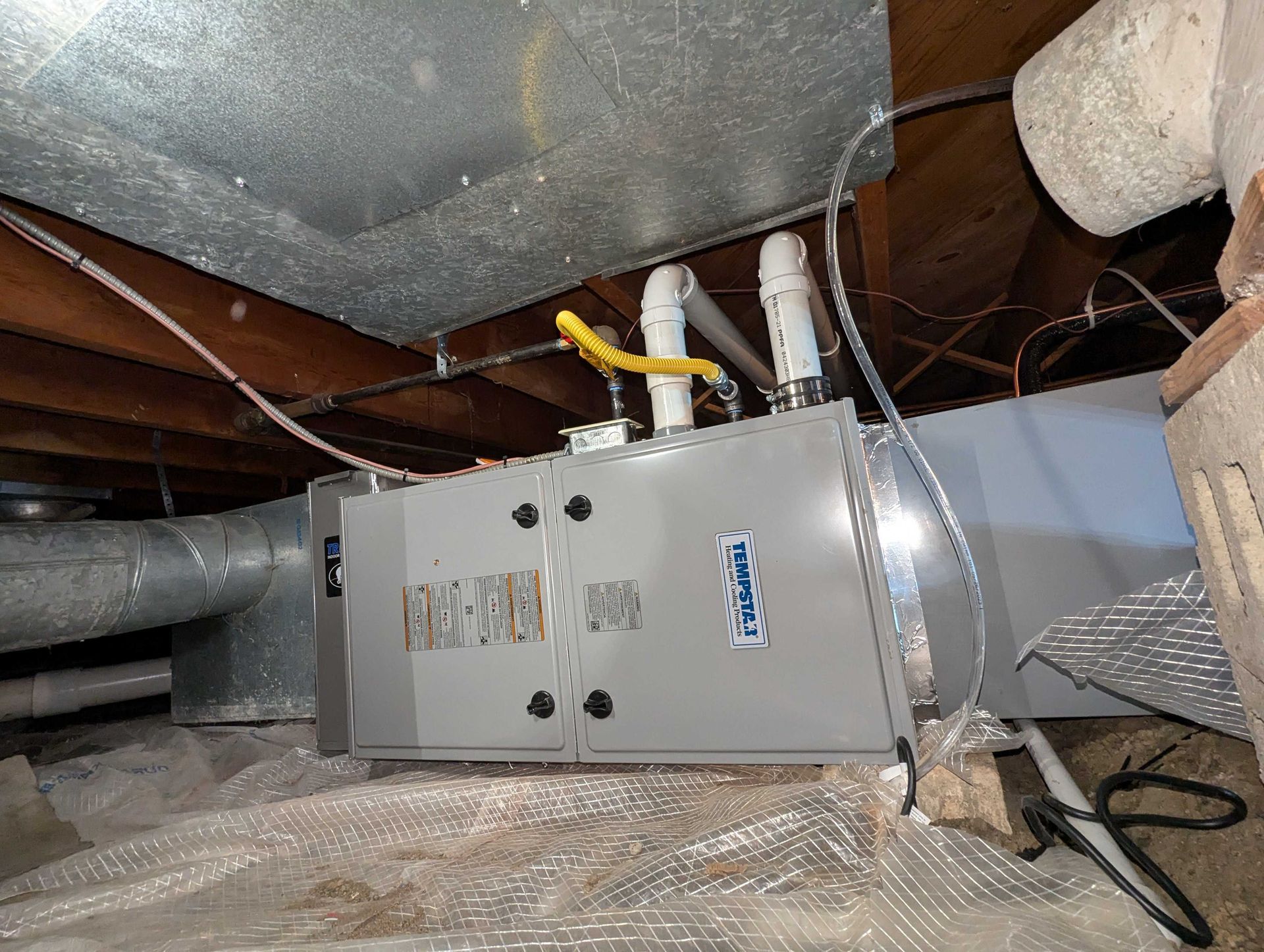 Furnace unit installed in a crawl space, with silver metal ductwork and gas lines visible.