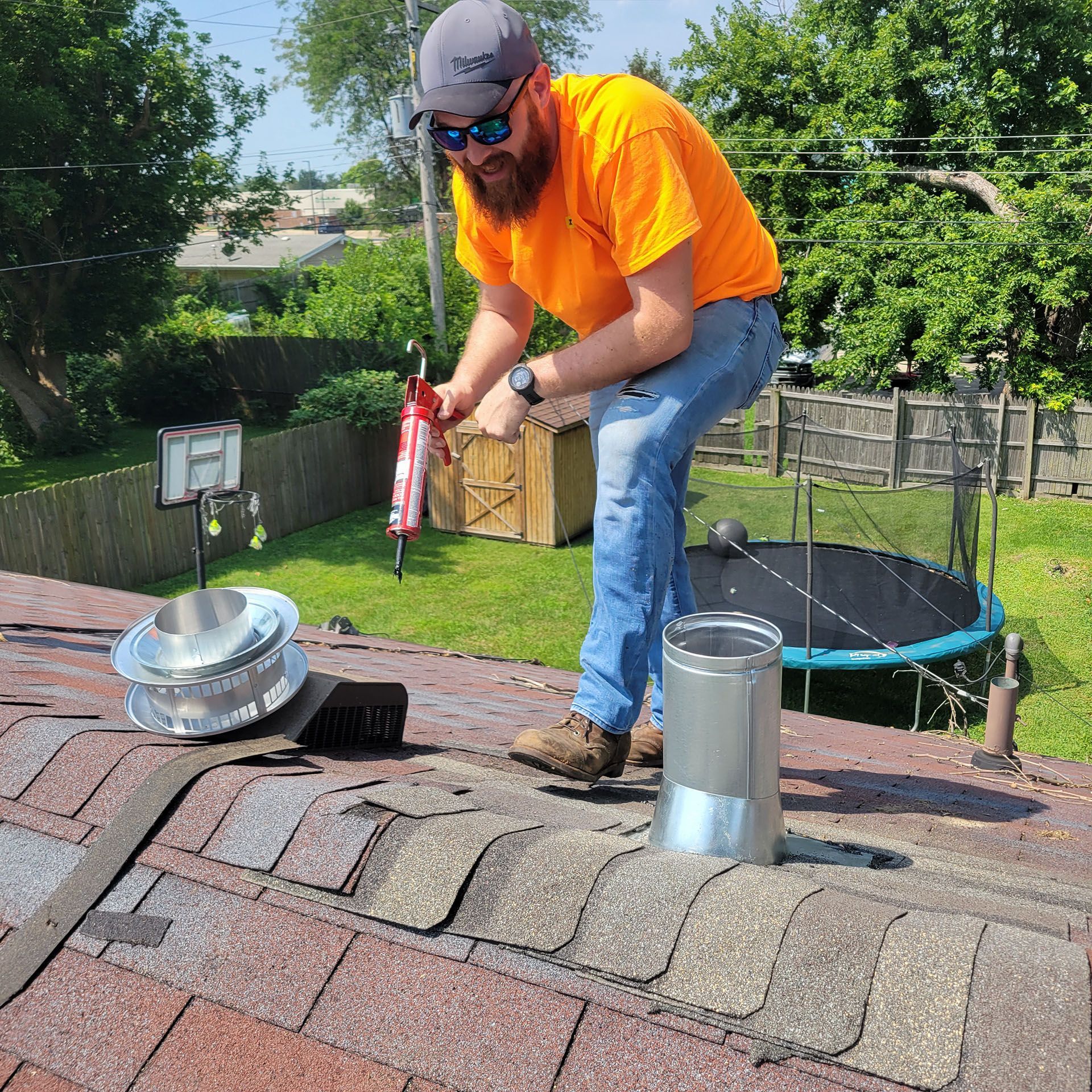 Man on a roof, applying sealant. He wears an orange shirt, jeans, and a hat. Chimney and parts visible.