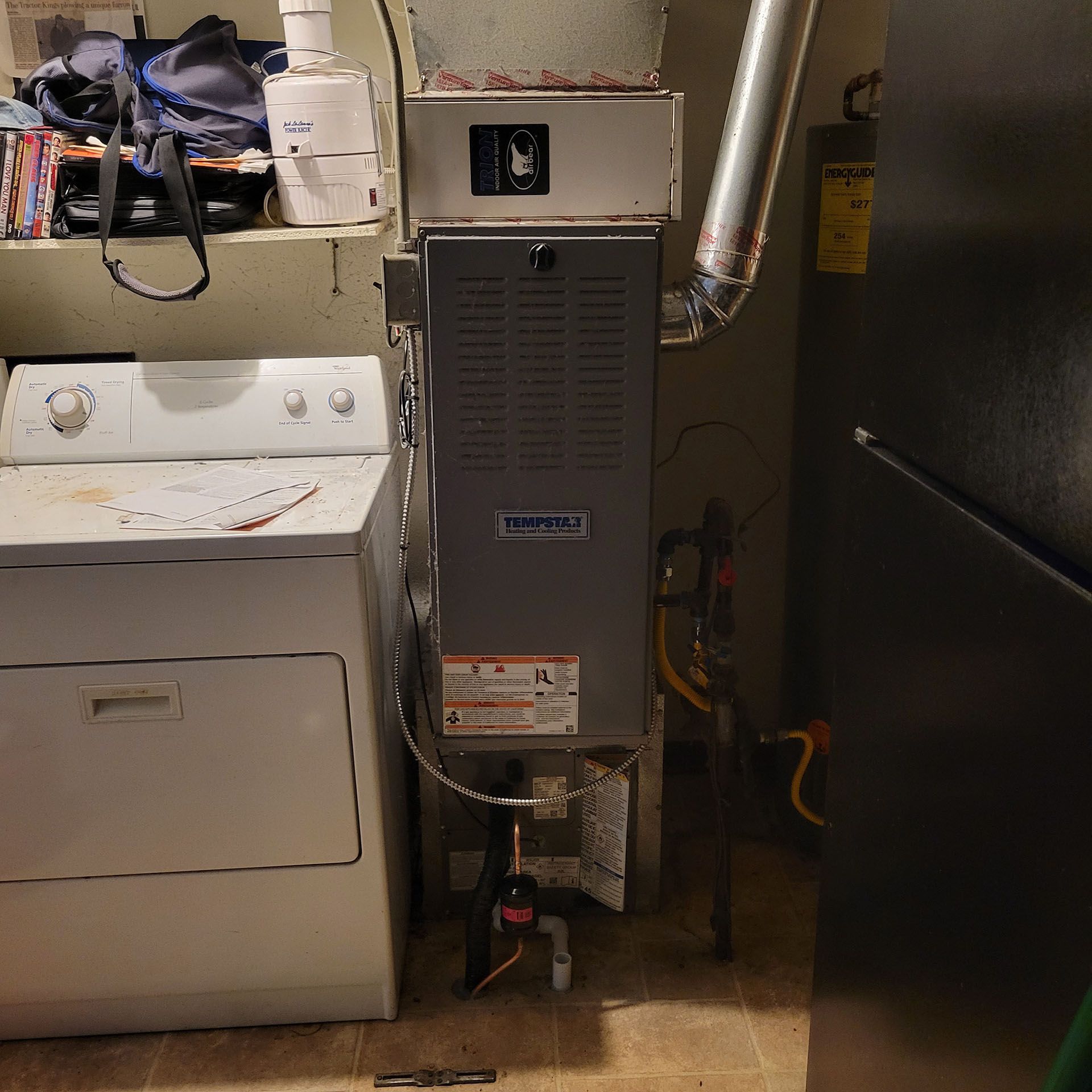 A furnace and dryer unit next to each other in a utility room with a shelf and refrigerator.