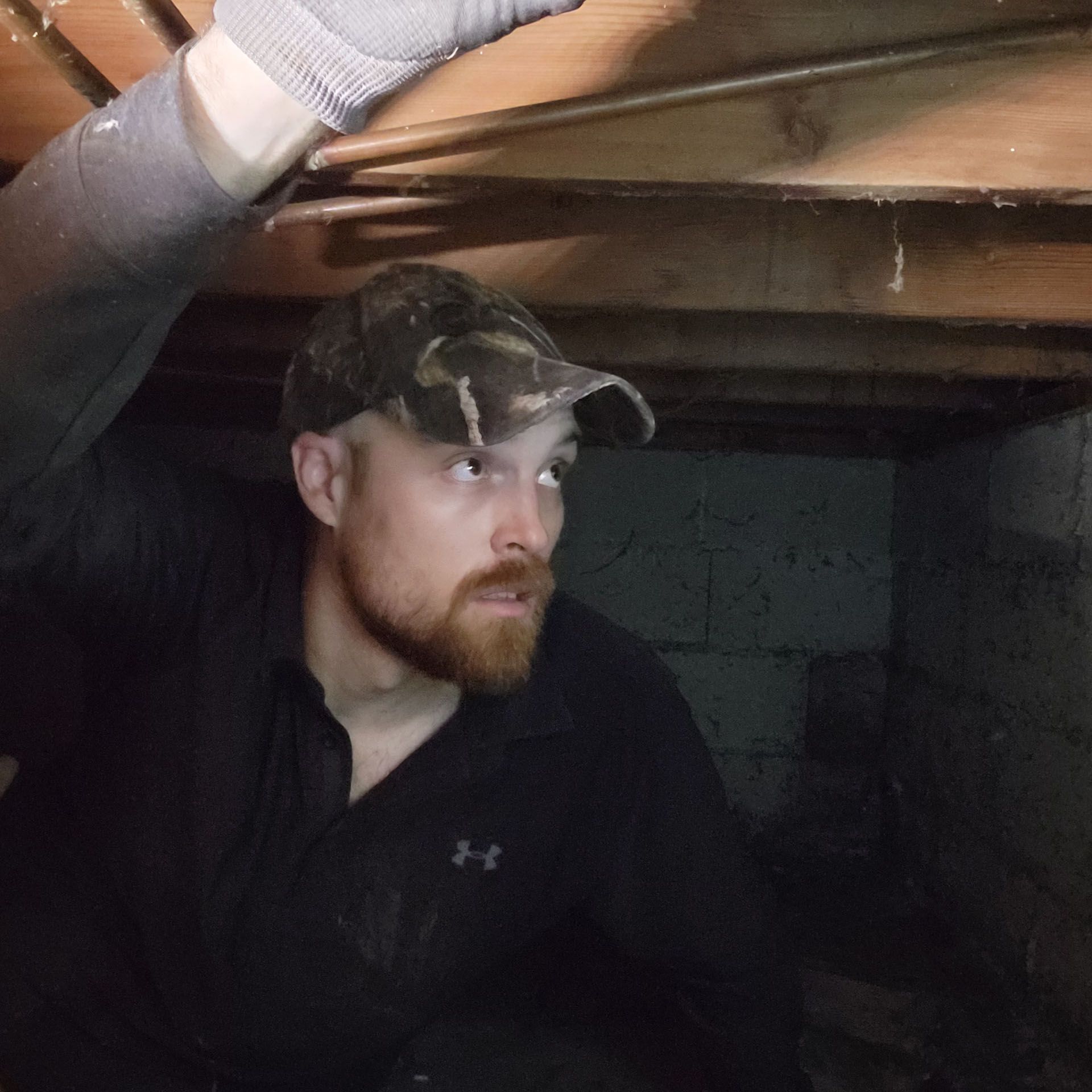 Man in a crawl space, wearing a hat and looking up with a focused expression.