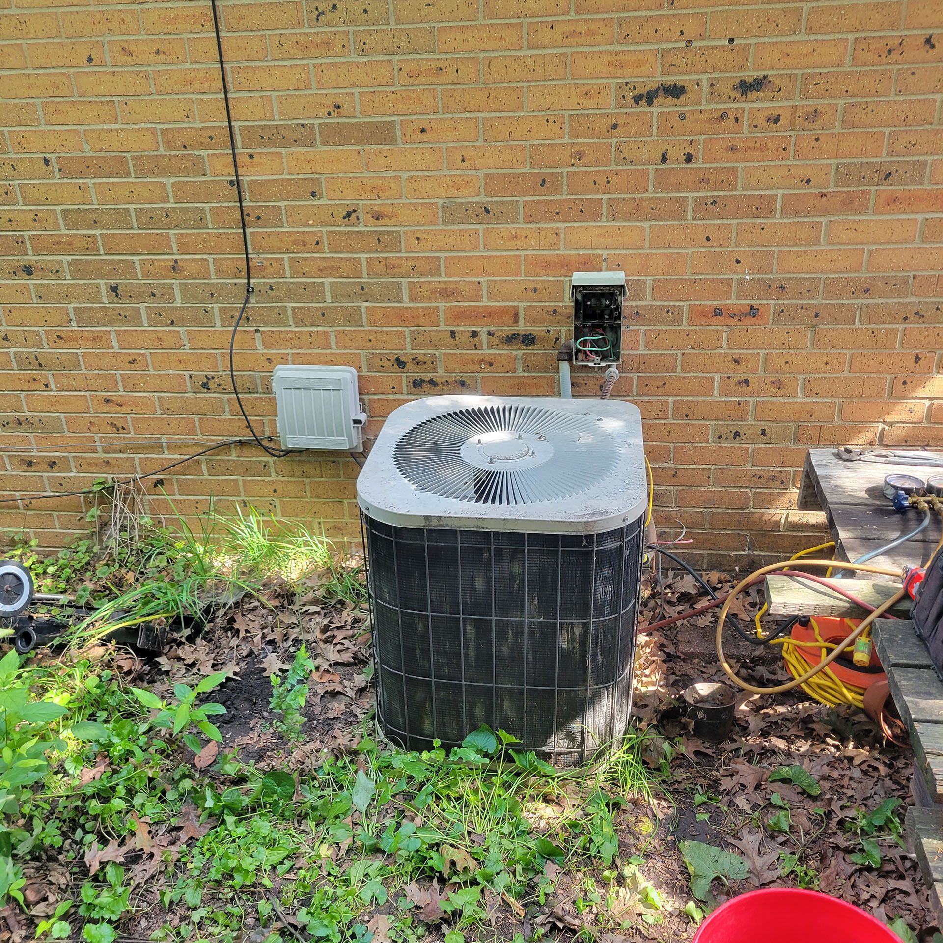 Air conditioning unit next to brick wall, surrounded by greenery and electrical components.